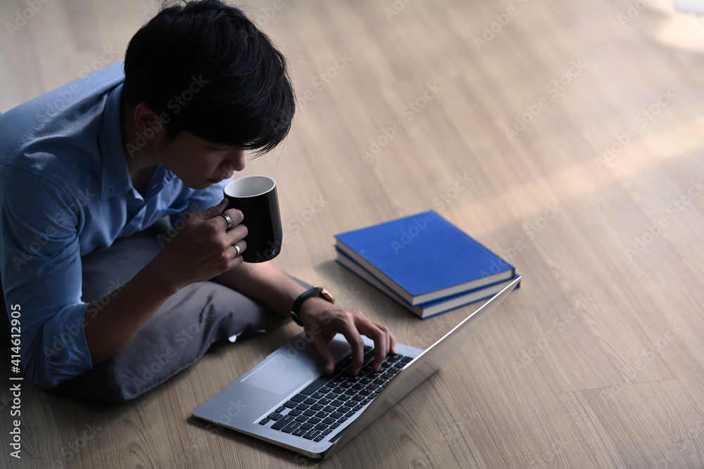 Young man working with computer laptop and drinking coffee while laying on wooden floor at home. Young man working with computer laptop and drinking coffee while laying on wooden floor at home.