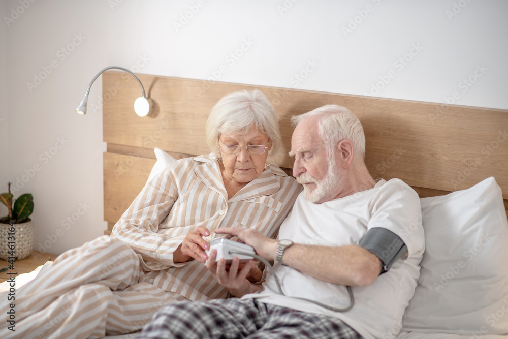 Grey-haired elderly man checking his blood pressure together with his wife Grey-haired elderly man checking his blood pressure together with his wife