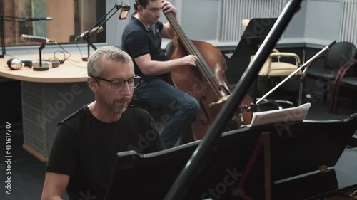 Medium handheld footage of concentrated male caucasian jazz pianist and contrabass player performing instrumental music at professional recording studio full of musical equipment