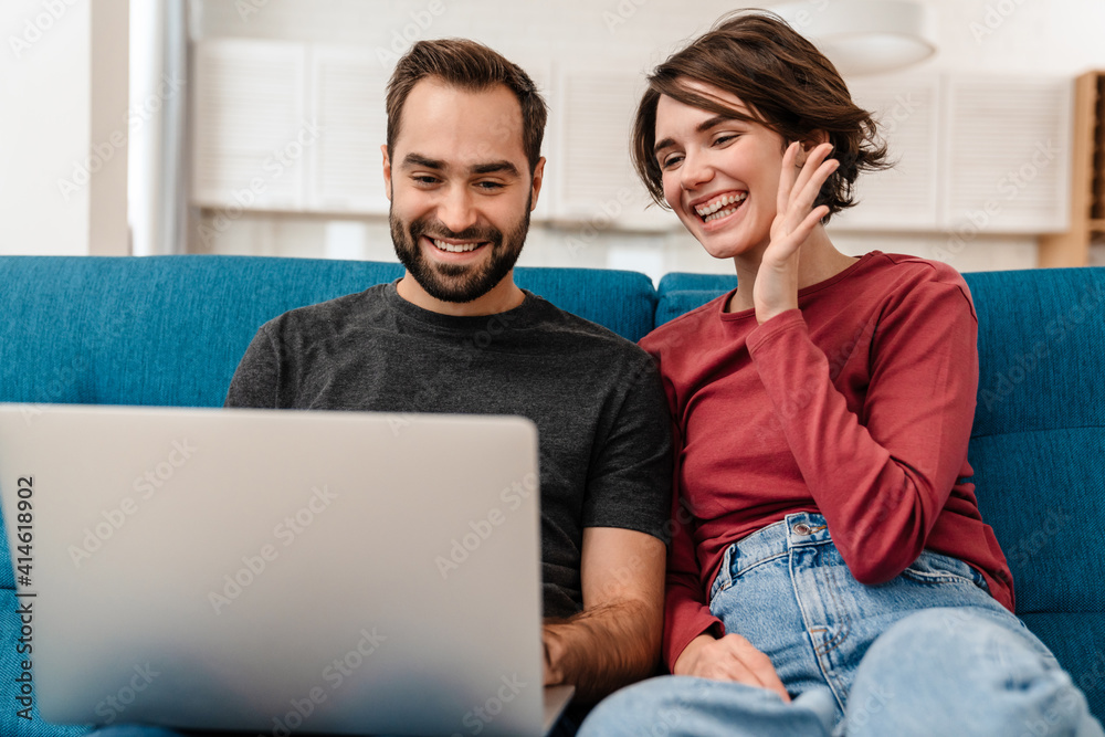 Happy young couple waving hand and using laptop while sitting on couch Happy young couple waving hand and using laptop while sitting on couch