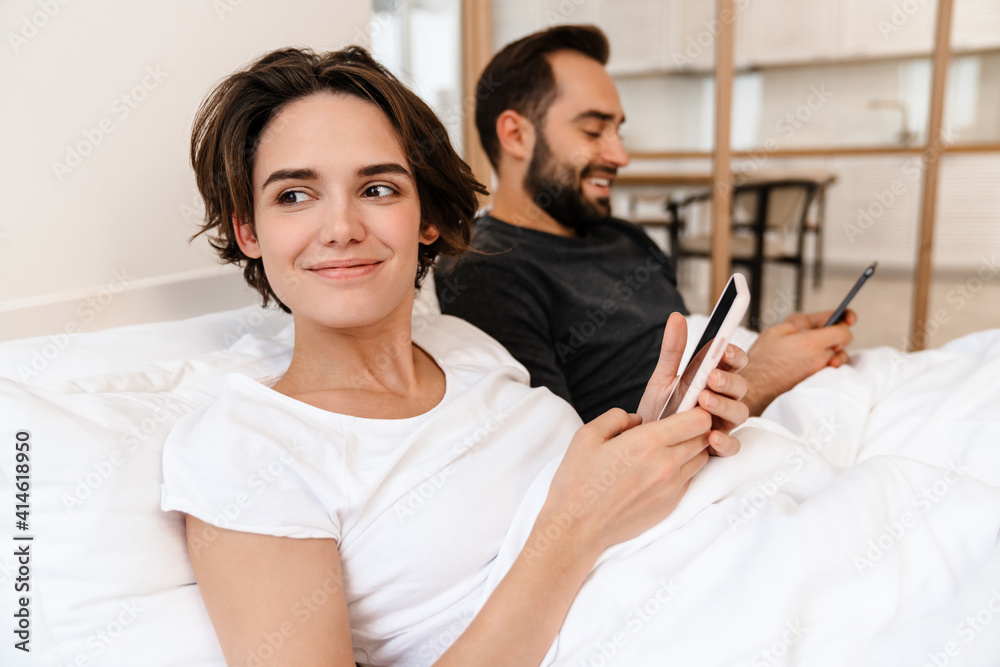 Happy young couple smiling and using mobile phones while lying in bed