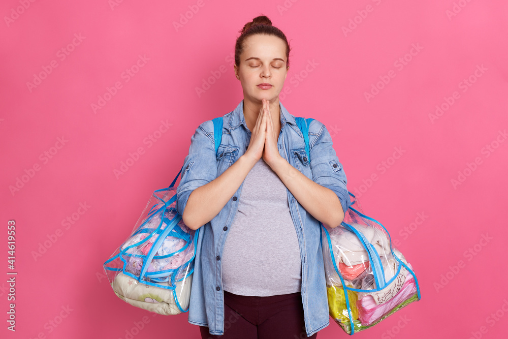 Beautiful pregnant woman with hair bun,expecting baby, keeps palms together and prays, posing isolated on pink background, wearing casual clothing, holding bags with stuff for maternity house. Beautiful pregnant woman with hair bun,expecting baby, keeps palms together and prays, posing isolated on pink background, wearing casual clothing, holding bags with stuff for maternity house.
