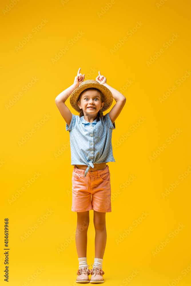 full length of happy child in straw hat pointing up with fingers on yellow