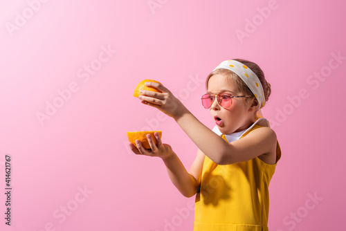 surprised girl in headscarf and sunglasses holding orange halves isolated on pink