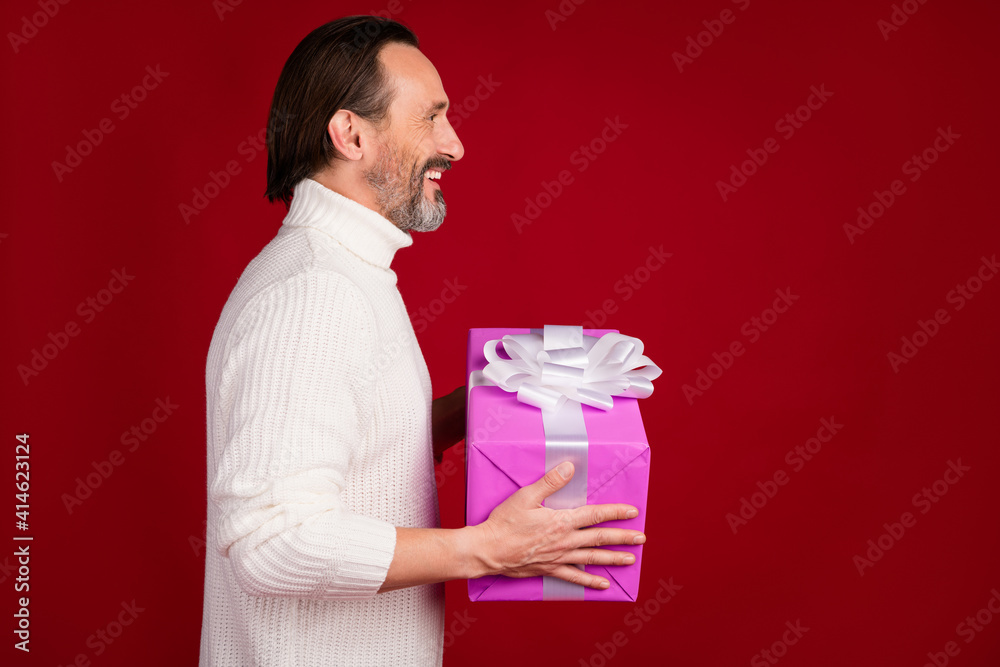 Profile side photo of mature happy positive good mod cheerful man gift present in copyspace isolated on red color background Profile side photo of mature happy positive good mod cheerful man gift present in copyspace isolated on red color background
