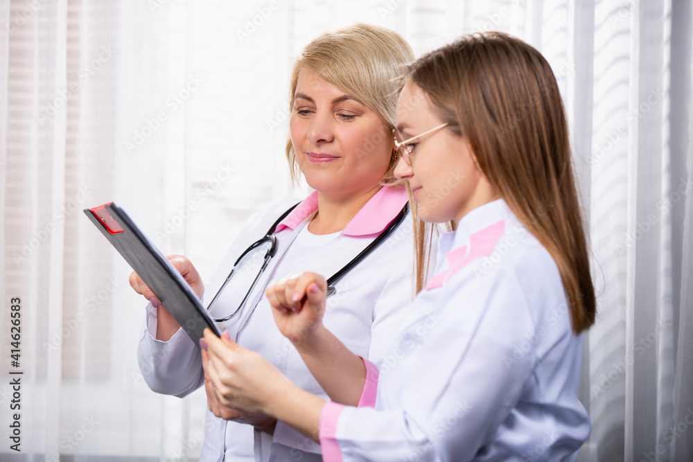 Profile photo of two medical workers discussing assignment sheet. Blond senior doctor, medical chief in white robe showing the assignment sheet to young medical assistant Profile photo of two medical workers discussing assignment sheet. Blond senior doctor, medical chief in white robe showing the assignment sheet to young medical assistant