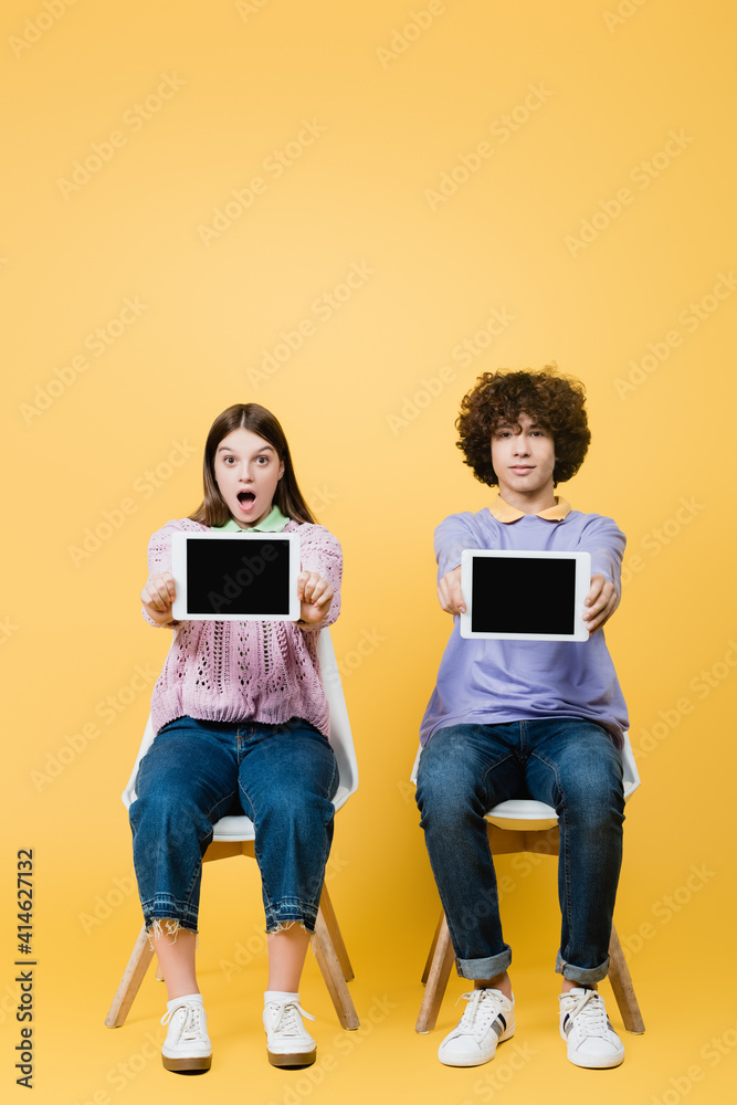 Excited girl showing digital tablet with blank screen near friend on chair on yellow background