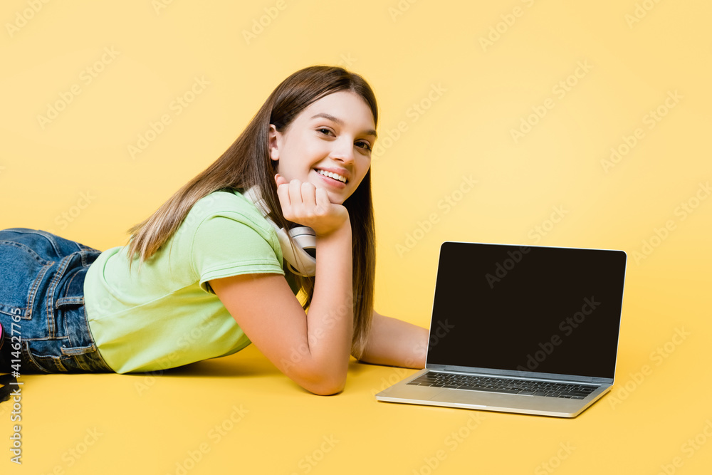 Smiling teenager in headphones looking at camera near laptop with blank screen on yellow background Smiling teenager in headphones looking at camera near laptop with blank screen on yellow background