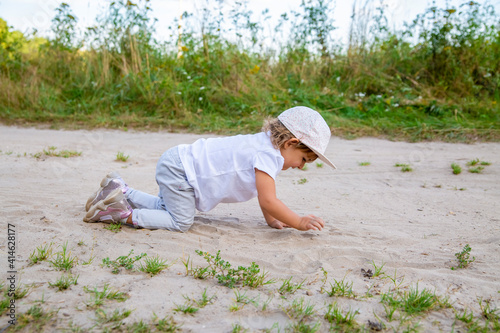 cute happy toddler in a cap crawls on all fours along a country road in the sand on a sunny summer day