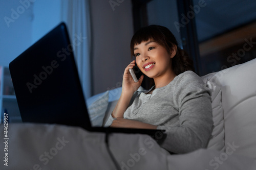 technology, internet, communication and people concept - happy smiling young asian woman with laptop computer calling on smartphone in bed at home at night