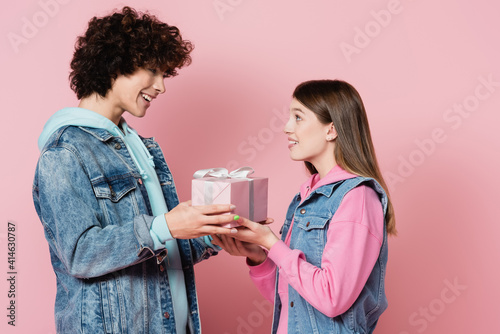 Smiling teen couple holding present with ribbon on pink background