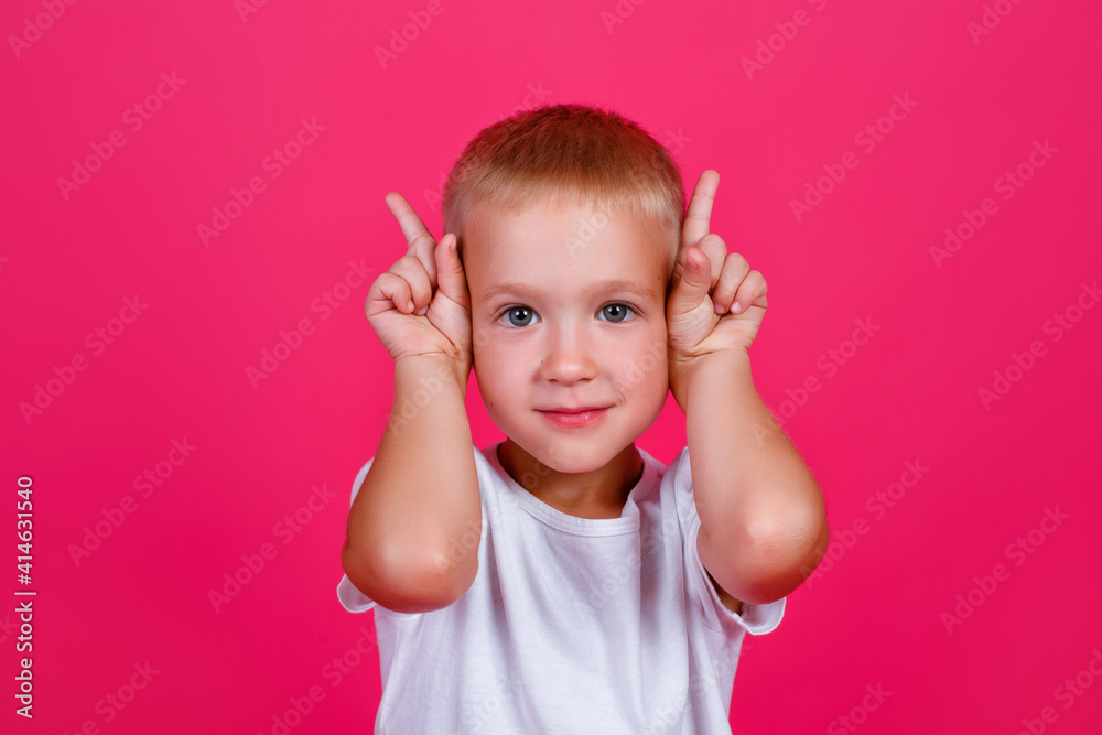 A portrait of a shocked blond boy in white casual clothes stands on a pink background, looking at the camera and pointing up with his index fingers. The surprised boy points to the copy space. A portrait of a shocked blond boy in white casual clothes stands on a pink background, looking at the camera and pointing up with his index fingers. The surprised boy points to the copy space.