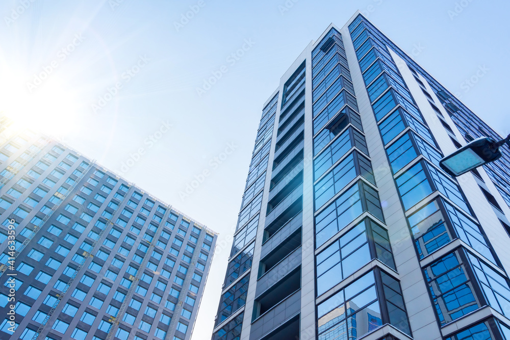 Multi-storey buildings with a glass facade and under construction with a facade department, with a bright sun in the sky.