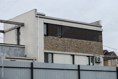 gray brown facade of a modern private house with small windows behind a metal fence against the sky