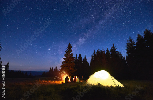 Evening camping near fire, spruce forest and mountains on background. Group of friends having a rest near bright bonfire. People sitting near tourist tent under night sky full of stars and Milky way.