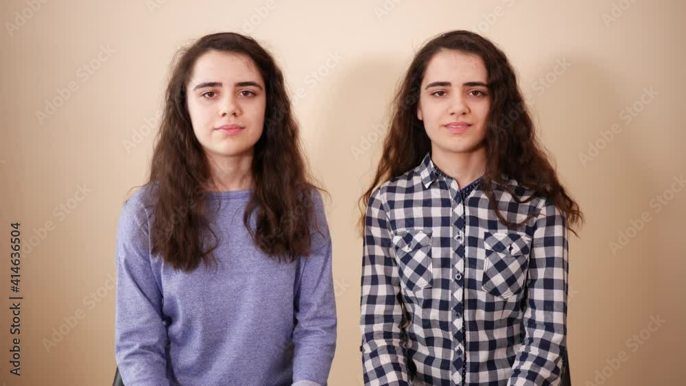 Two twins sisters teenage cute girls with curly hairs sit along watch to camera posing at home in a room Two twins sisters teenage cute girls with curly hairs sit along watch to camera posing at home in a room