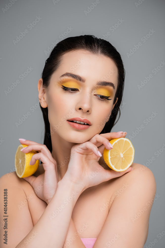 brunette woman with bare shoulders holding yellow lemon halves isolated on grey
