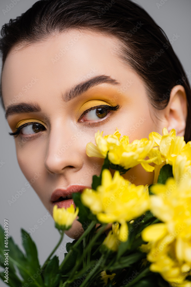 young brunette woman with makeup near blooming yellow flowers on blurred foreground isolated on grey young brunette woman with makeup near blooming yellow flowers on blurred foreground isolated on grey