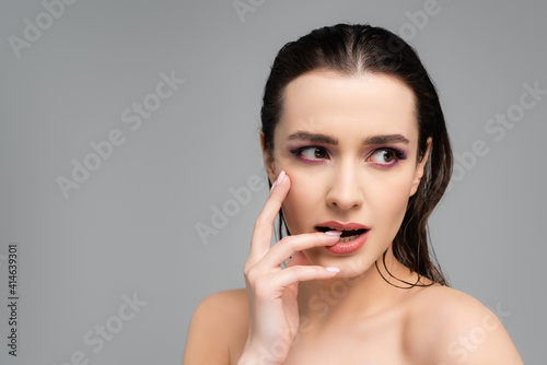 young woman with pink eye shadows and bare shoulders biting nail isolated on grey