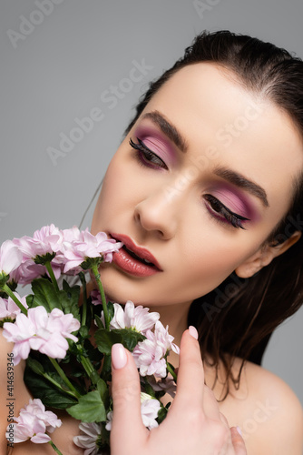 young woman with pink eye shadows looking away near flowers isolated on grey