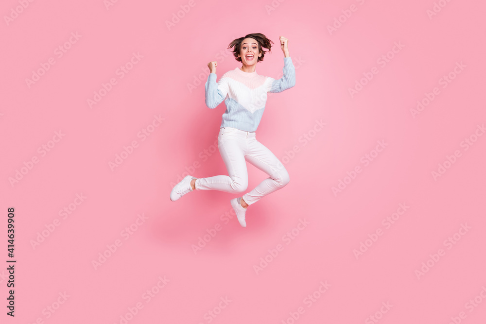 Full length photo of funky lucky young woman wear sweater jumping rising fists isolated pink color background