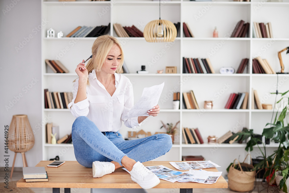 Young blond woman, wearing casual jeans and white top, sitting on table, working on design project. Freelancer employed at home. Work process in modern company office. Young successful professionals. Young blond woman, wearing casual jeans and white top, sitting on table, working on design project. Freelancer employed at home. Work process in modern company office. Young successful professionals.