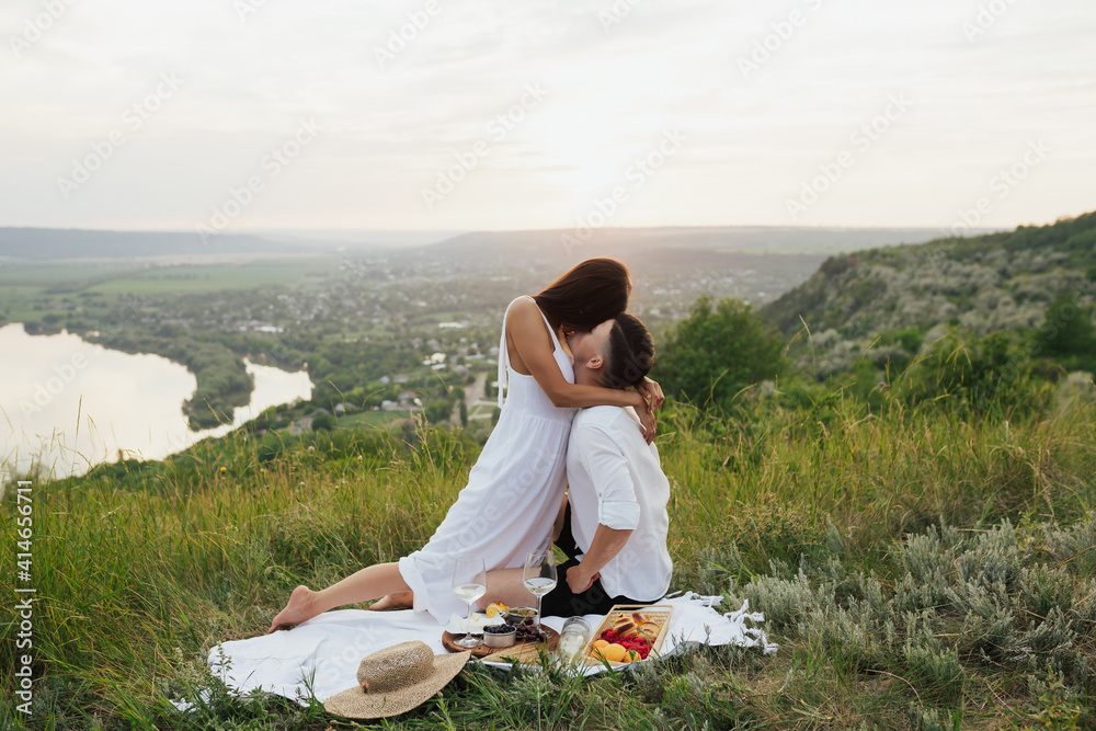 Passionate couple in love hugging and kissing on a summer picnic while sitting on the hill with beautiful landscape on background.
