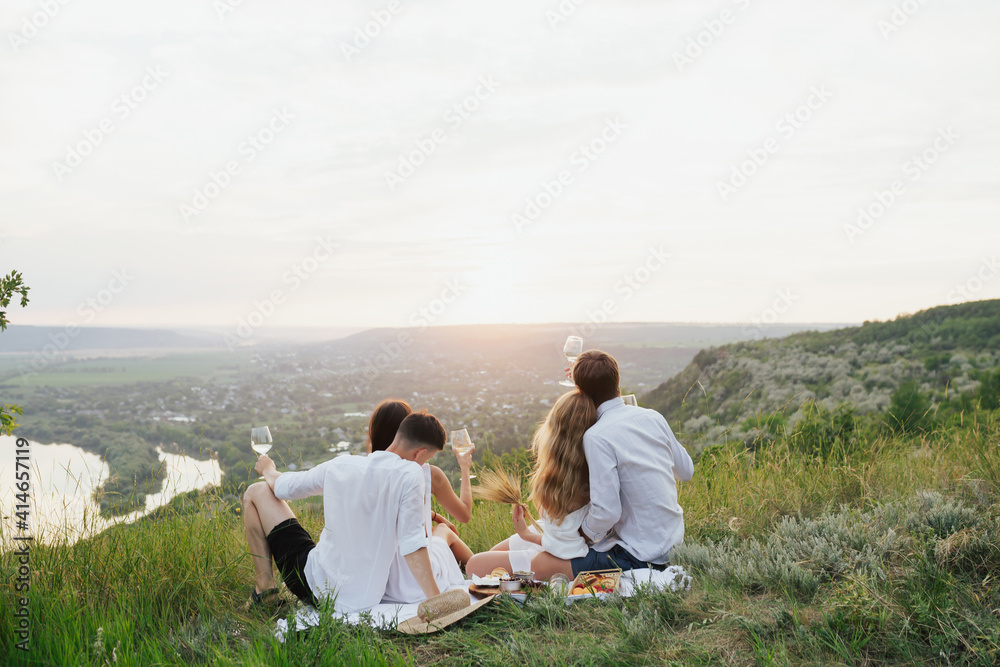 Group of friends is having a picnic on the hill with beautiful landscape on background. Young people sit together on the grass. 
