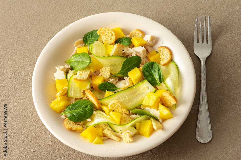 Plate with salad and fork close-up on a brown background, salad of cucumber and chicken breast, mozzarella and croutons with sweet mango pieces and spinach