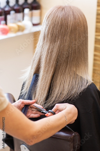 Close-up of the hands of a professional hair stylist with scissors and comb. Repairing long hair of a blonde woman. Process of cutting split ends. Haircut at a beauty salon. Woman's hairstyle.