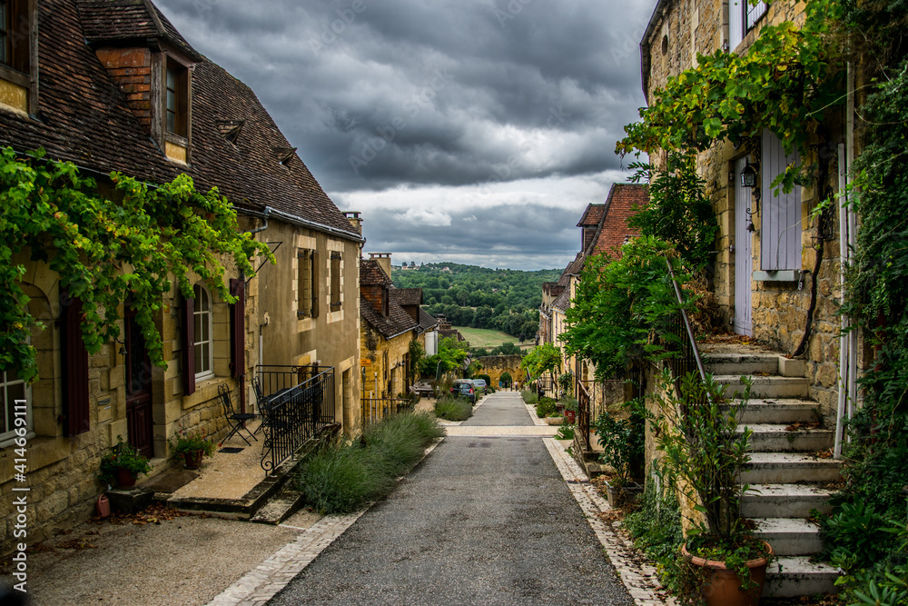 Calle que desciende hacia las murallas y el campo, con casas de piedra con ventanas y vegetación en sus fachadas en una aldea histórica medieval francesa