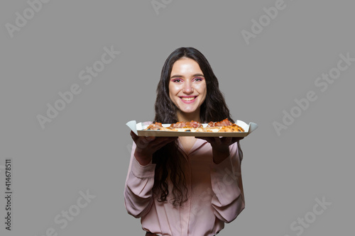 Girl courier with pinsa romana gourmet italian cuisine on grey background. Holding scrocchiarella traditional dish. Food delivery from pizzeria. Pinsa with meat  arugula  olives  cheese.