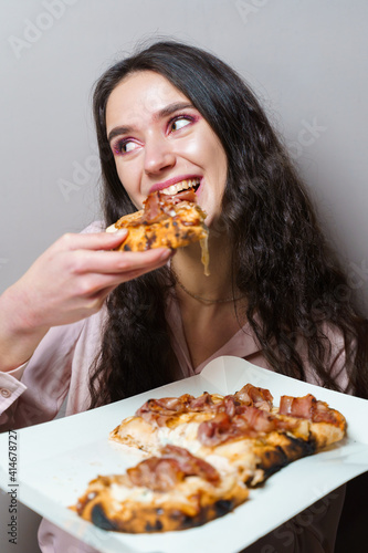 Girl courier eating pinsa pizza romana gourmet italian cuisine on grey background. Holding scrocchiarella traditional dish. Food delivery from pizzeria. Pinsa with meat  arugula  olives  cheese.