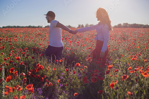Handsome young guy andbeautiful  girl walking on the field of poppies on a beautiful warm sunset. Girl leads the boy holding the hand photo