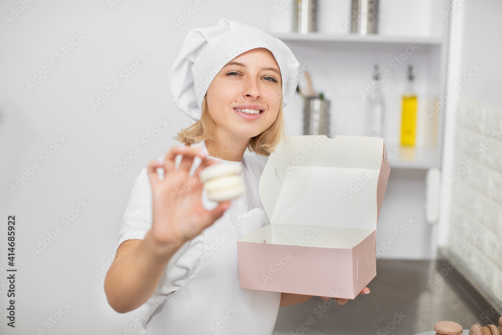 Close up portrait of pretty blond woman pastry chef, wearing white hat and apron, demonstrating to camera tasty white macarons. Focus on the woman's face