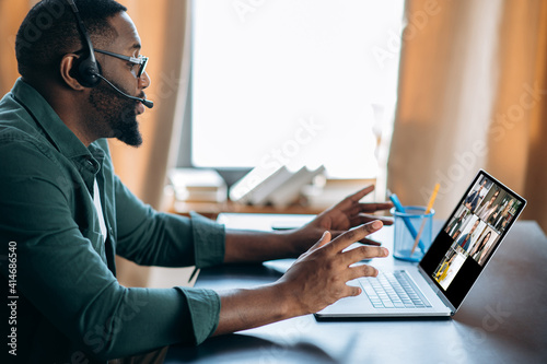 Virtual conference with multi ethnic people. African American guy in headset learns remotely using laptop and video chatting app. On the laptop screen the teacher and group of diverse people, online photo