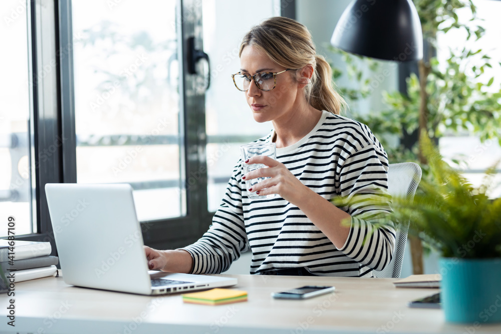Beautiful mature business woman working with a laptop while drinking glass of water on a desk in the office at home. Beautiful mature business woman working with a laptop while drinking glass of water on a desk in the office at home.