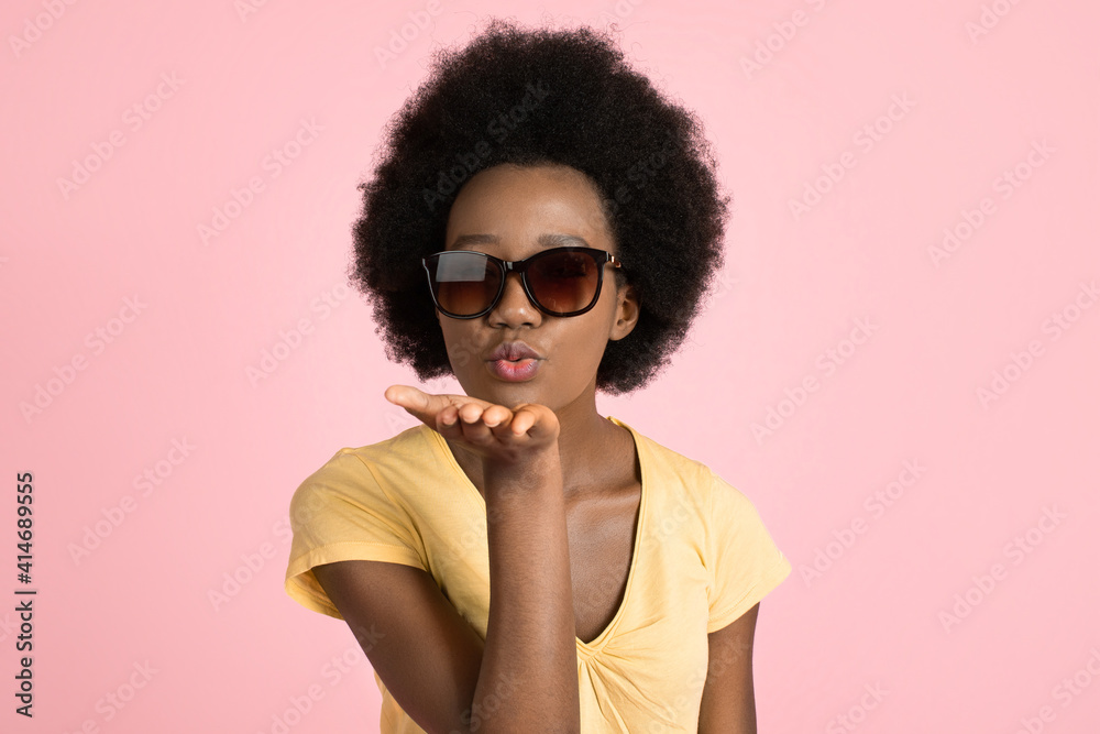 Sunny bright portrait of young black African woman with afro hair style, wearing sunglasses and yellow t-shirt, making air kiss to camera, posing over isolated pink background