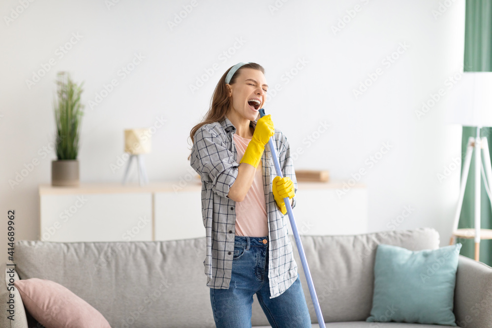 House cleaning is fun. Cheerful young lady using mop as mic, singing song while doing cleanup at living room House cleaning is fun. Cheerful young lady using mop as mic, singing song while doing cleanup at living room