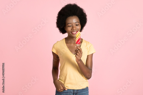 Horizontal studio shot of attractive young African American woman with afro hair style  wearing yellow t-shirt  standing on isolated pink background and looking at lollipop in hand