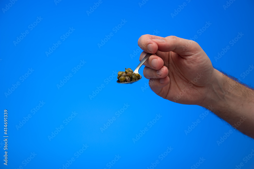 Caucasian male hand holding a spoon full of pickled capers isolated on blue background studio shot Caucasian male hand holding a spoon full of pickled capers isolated on blue background studio shot