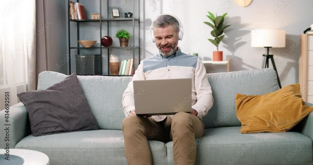 Cheerful Caucasian middle-aged bearded man sitting on sofa in cozy room in headphones watching comedy movie or videos on laptop computer and laughing, positive emotions, leisure concept Cheerful Caucasian middle-aged bearded man sitting on sofa in cozy room in headphones watching comedy movie or videos on laptop computer and laughing, positive emotions, leisure concept
