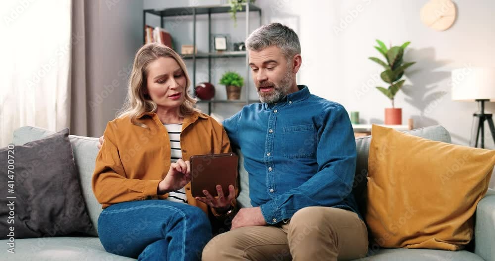 Portrait of cute Caucasian middle-aged married couple sitting on sofa in living room searching internet on tablet device browsing online and choosing something, e-commerce, leisure concept