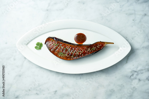 Baked eggplant in josper with miso sauce, cilantro, nuts and sesame seeds. Miso eggplant in a white plate on a marble background. View from above photo