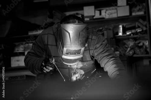 An adult male works in an old workshop. He holds a welding machine in his hand and welds the metal structure. Black and white.