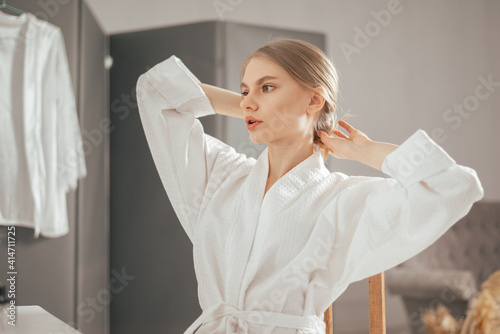 Young blonde beautiful woman in the white bathrobe looking to the mirror and touching her hair