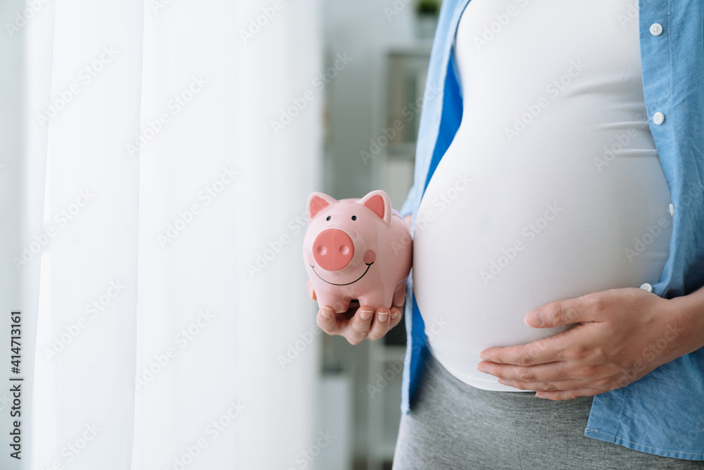 close up unrecognized asian pregnant woman holding pink piggy bank and  keeping one hand on big belly. focus view on future mom abdomen with coin  bank. maternity motherhood saving money concept. Photos