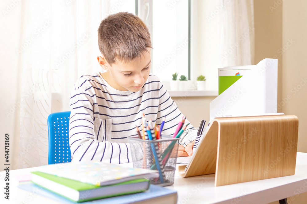 Boy with digital tablet computer writing on notebook doing homework at table. Online learning, remote education, distance lessons at home Boy with digital tablet computer writing on notebook doing homework at table. Online learning, remote education, distance lessons at home