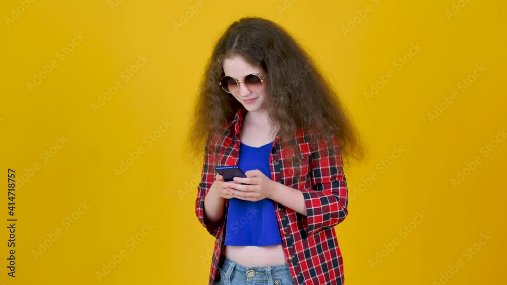 Young happy girl with curly hair wearing glasses using smart phone and texting online chat with smile isolated on yellow background. People lifestyle concept. Young happy girl with curly hair wearing glasses using smart phone and texting online chat with smile isolated on yellow background. People lifestyle concept.