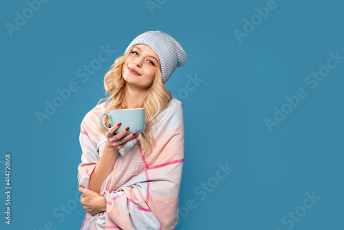 Beautiful smiling blonde woman in winter fashionable cap and scarf smiling to the camera, drinking hot tea, isolated on blue studio background. Real people emotions. A lot of copy space.